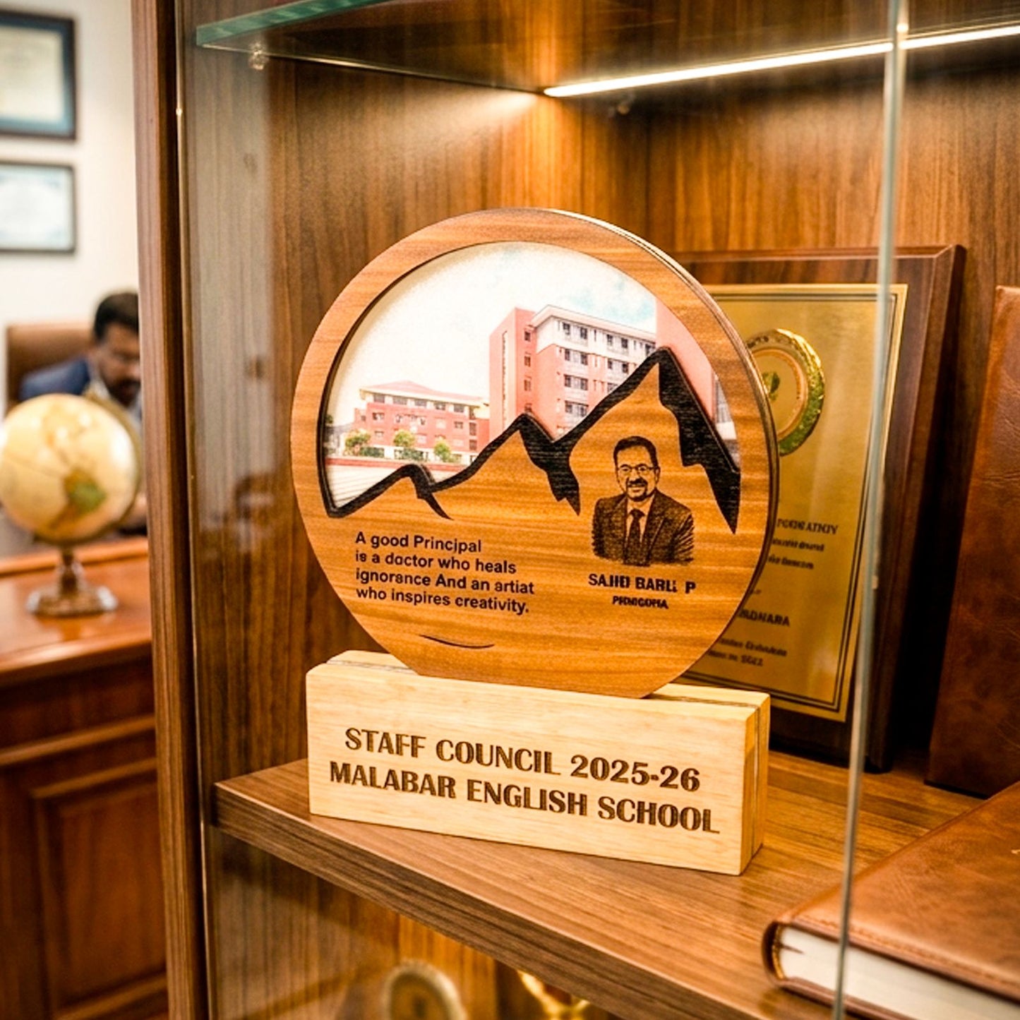 A round wooden school award plaque displayed on a wooden stand inside a glass cabinet. The plaque features an engraved mountain and portrait of media